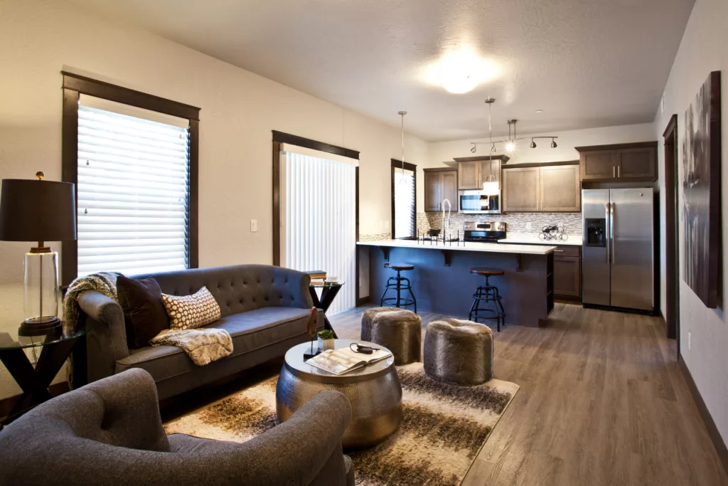Living room and galley kitchen. Stainless steel appliances, two bar stools, couch in front of window, chair ottoman.